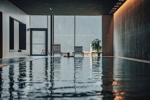 An indoor swimming pool with clear glass walls, two chairs, and a potted plant.