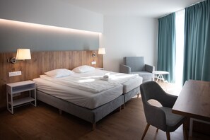 Ein Hotelzimmer mit Doppelbett, zwei Nachttischen, einem Sessel, einem kleinen runden Tisch und einem Fenster mit Vorhängen.