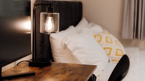 Een net opgemaakt bed met kussens in wit en geel dessin, een zwart hoofdbord en een nachtkastje met een moderne lamp.