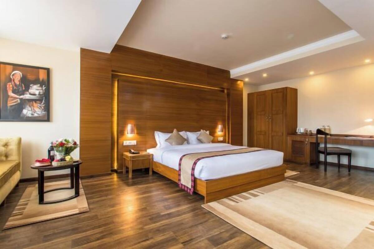 A modern bedroom with a wooden headboard, a bed with white linens, a nightstand with a lamp, a small table with flowers, and a painting on the wall.