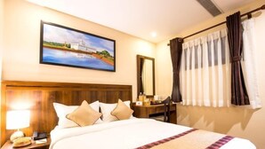 A hotel room with a large bed, a nightstand with a lamp, a desk with a chair, a painting on the wall, and a window with curtains. - Hotel Kutumba (Lalitpur)