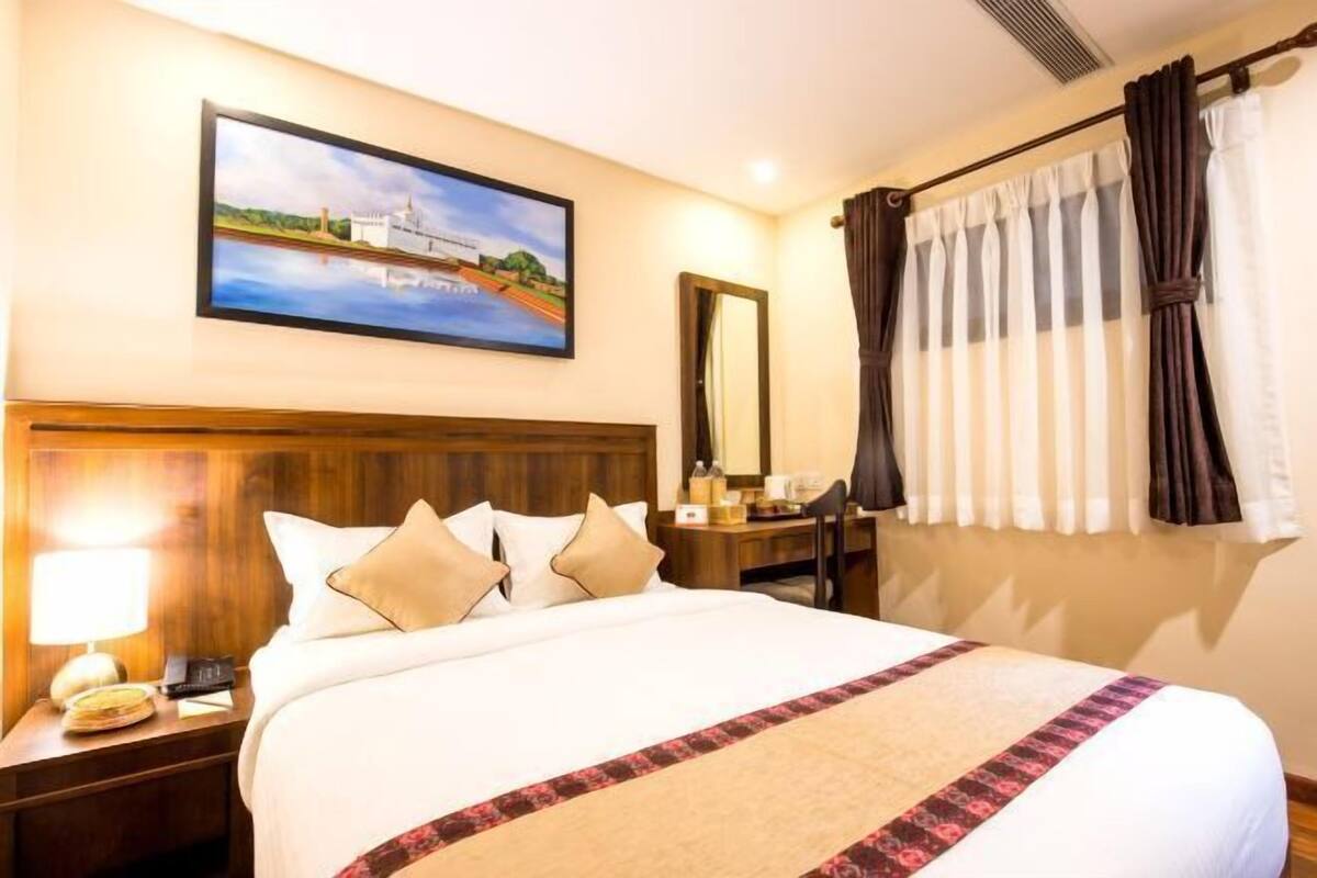 A hotel room with a large bed, a nightstand with a lamp, a desk with a chair, a painting on the wall, and a window with curtains.