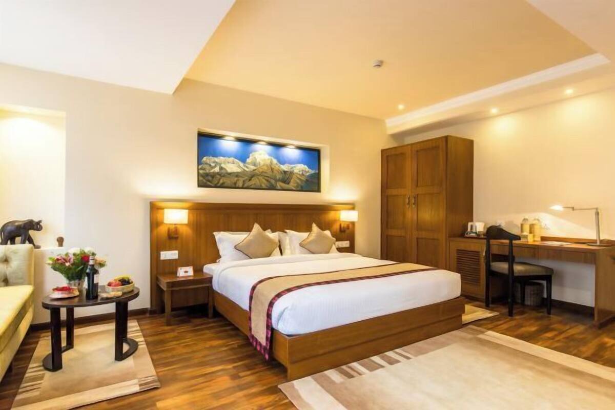 A hotel room with a bed, bedside table, lamp, desk, chair, and a painting on the wall.