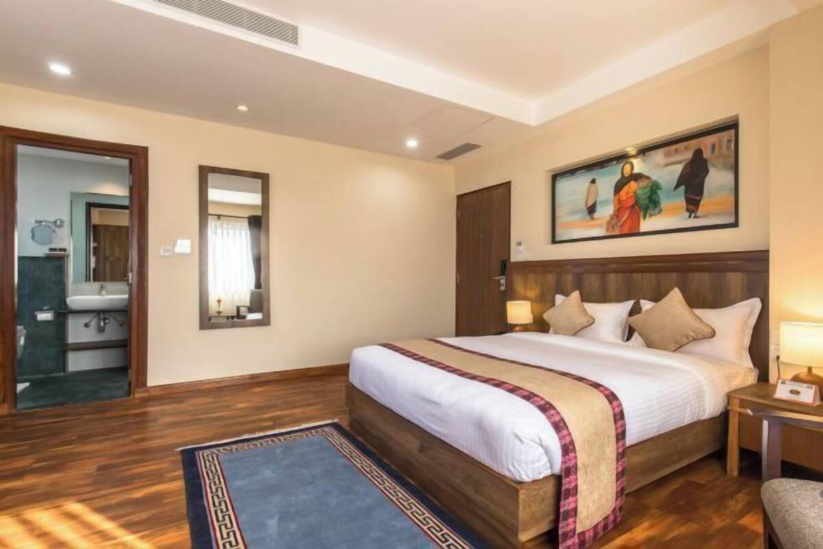 A hotel room with a large bed, a wooden headboard, a painting on the wall, a bedside table with a lamp, and a bathroom visible through an open door.