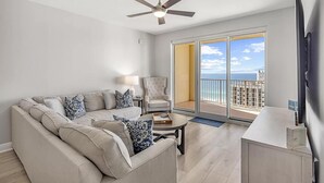 A living room with a sofa, armchair, coffee table, and a ceiling fan. There is a sliding glass door leading to a balcony with a view of the ocean.