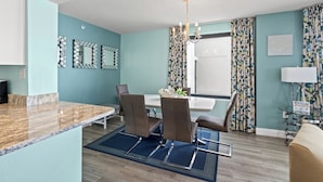 Una cocina moderna con encimera de mármol, zona de comedor con dos sillas, candelabro y ventana con cortinas estampadas.