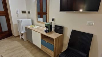 Un espace de vie compact comprenant un bureau en bois, une chaise noire, un téléviseur fixé au mur et un réfrigérateur.