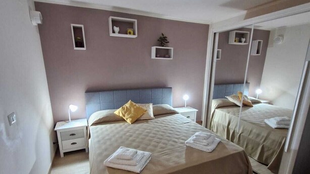 A bedroom with a large bed, bedside tables, a mirror, and a small shelf with decorative items. - Medea Appartamenti (Siracusa)