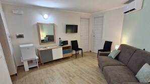 A compact living space with a brown sofa, a small desk with a mirror, a television, and a white wardrobe. - Medea Appartamenti (Siracusa)