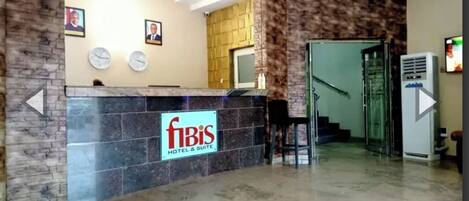 A hotel reception area with a reception desk, framed pictures on the wall, a staircase, and an air conditioning unit.