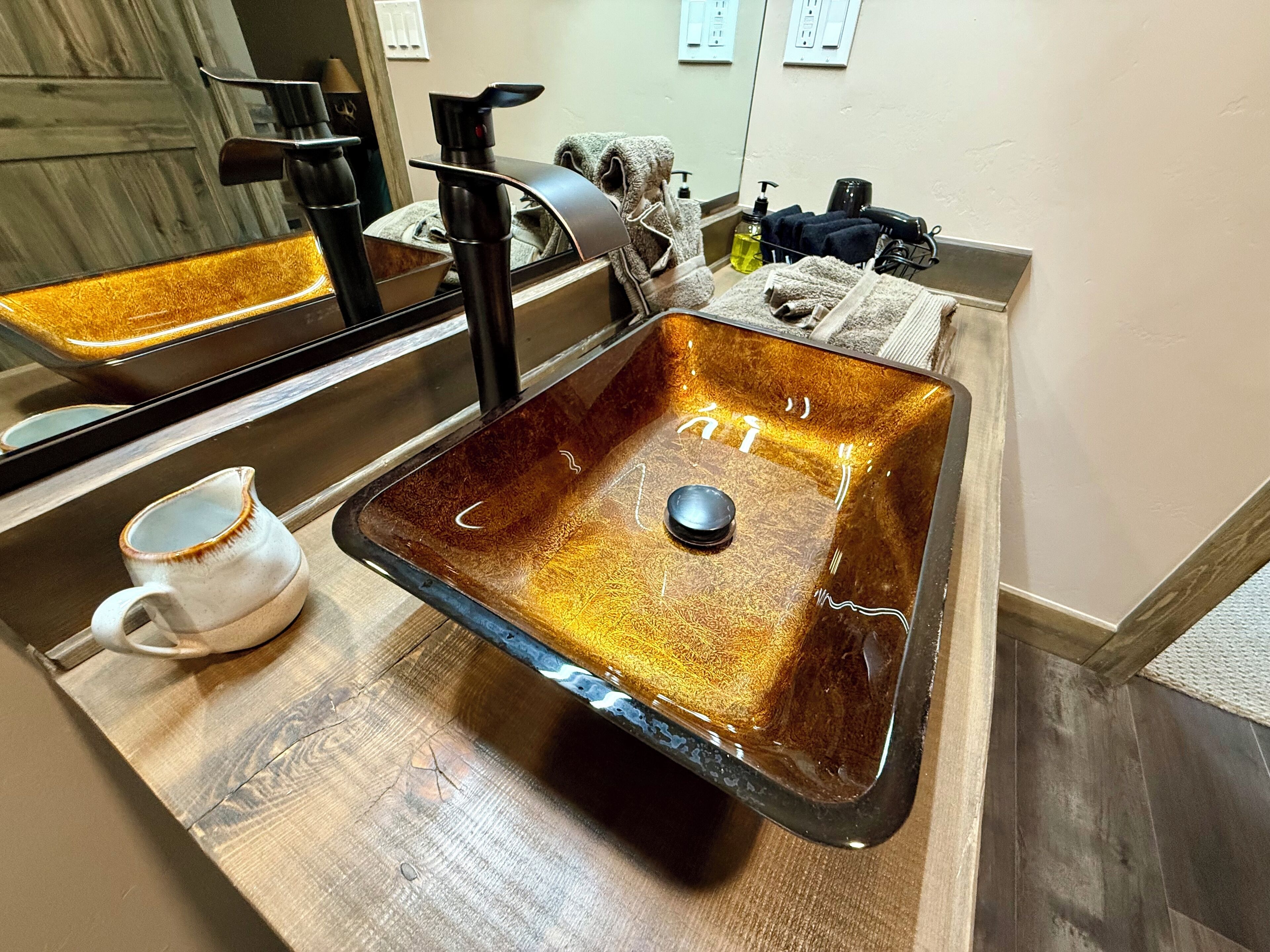 Un baño con un lavamanos rectangular, un espejo, un perchero para toallas y una taza sobre el mostrador.