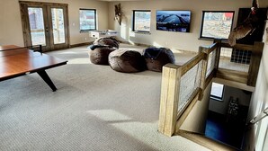 Una habitación amplia con mesa de ping pong, pufs, un televisor grande y vistas al exterior.