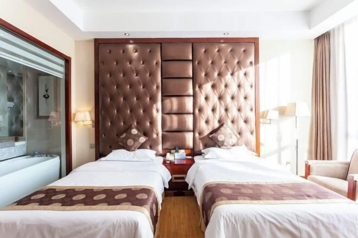 A hotel room with two beds, a large padded headboard, a glass shower enclosure, and a sitting area with a chair.