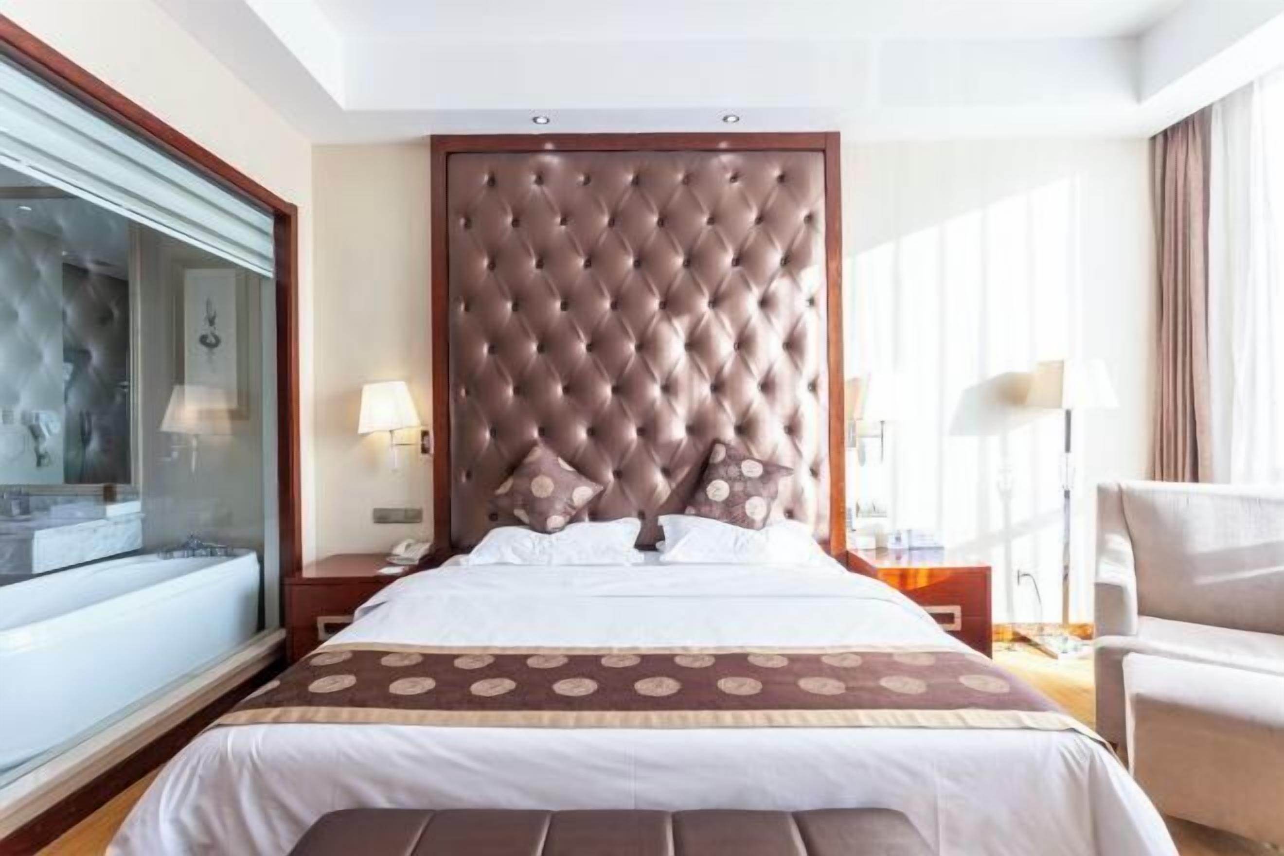 Habitación de hotel con una cama grande, un baño con paredes de vidrio y un cabecero de madera, además de una zona de estar con una silla.
