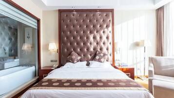 Habitación de hotel con una cama grande, un baño con paredes de vidrio y un cabecero de madera, además de una zona de estar con una silla.