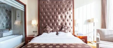 A hotel room with a large bed, a glass-enclosed bathroom, a wooden headboard, and a seating area with a chair.