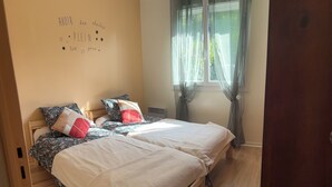 A bedroom with two beds, floral bedding, a wooden floor, and a window with sheer curtains.
