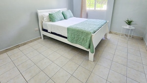 Una cama bien hecha con estructura blanca y ropa de cama verde, una pequeña mesa blanca con una planta, y una ventana con vistas a zonas verdes.