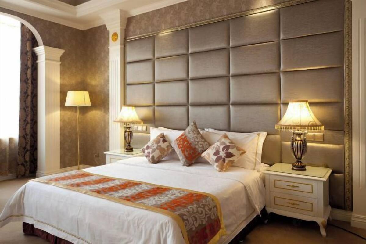 A hotel room with a large bed, two bedside lamps, a decorative headboard, and a patterned bedspread.