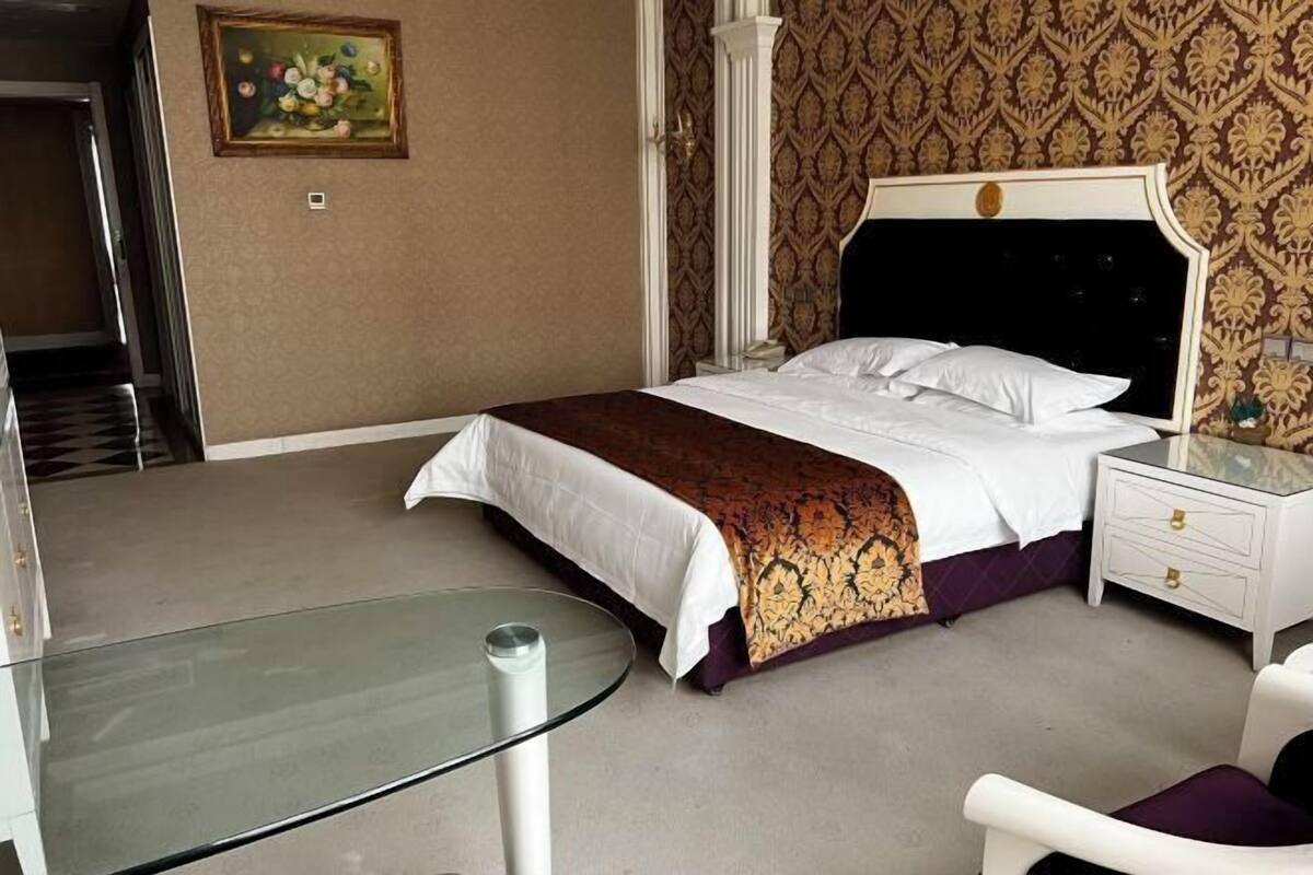 A hotel room with a bed, a glass table, a white nightstand, and a chair.