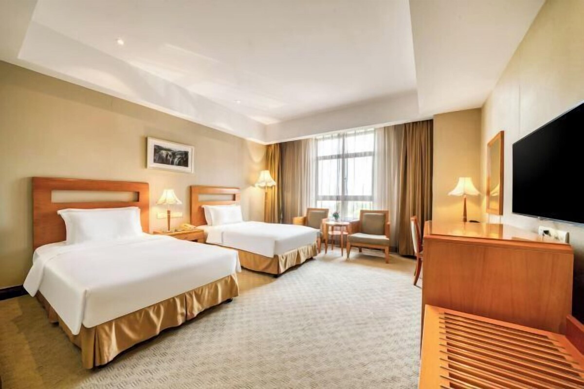 A hotel room with two beds, a desk, a TV, and a window with curtains.