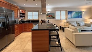 Una cocina moderna con una isla central, armarios de madera y un refrigerador de acero inoxidable. Hay un área de comedor con un sofá blanco y un cuadro en la pared.