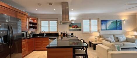 A modern kitchen with a central island, wooden cabinets, and a stainless steel refrigerator. There is a dining area with a white sofa and a painting on the wall.