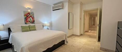 Un dormitorio con cama, ventilador de techo, equipo de aire acondicionado, un cuadro y un pasillo que conduce a otra habitación.