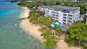 Vista aérea de un hotel frente a la playa con varios pisos, piscina y zona de playa con aguas turquesas cristalinas.