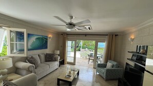 Una sala de estar con un sofá, sillones, una mesa de centro y un televisor. Hay un ventilador de techo, un cuadro y vistas al exterior a través de una puerta corrediza.