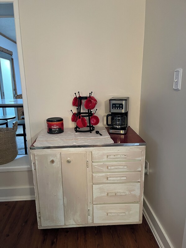 Una kitchenette con cafetera, un escurreplatos para tazas y un bote de café Folgers.
