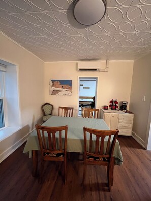 A dining area with a table and chairs, an air conditioning unit, a painting on the wall, and a kitchen area with cabinets and appliances.