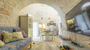 A cozy living room with a sofa, a dining table, and a TV. The room features stone walls and an arched ceiling.