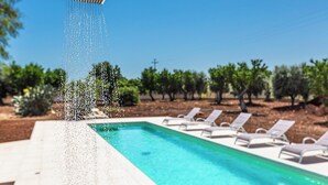 A swimming pool with water cascading from a showerhead, surrounded by lounge chairs and a backdrop of trees.