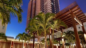 A modern hotel with high-rise buildings in the background, palm trees, and a wooden pergola structure.