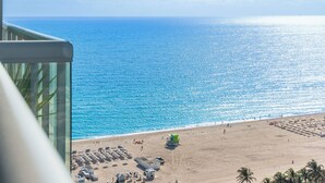 A beach view from a balcony with a clear blue sea, a sandy beach with umbrellas, and a few people.