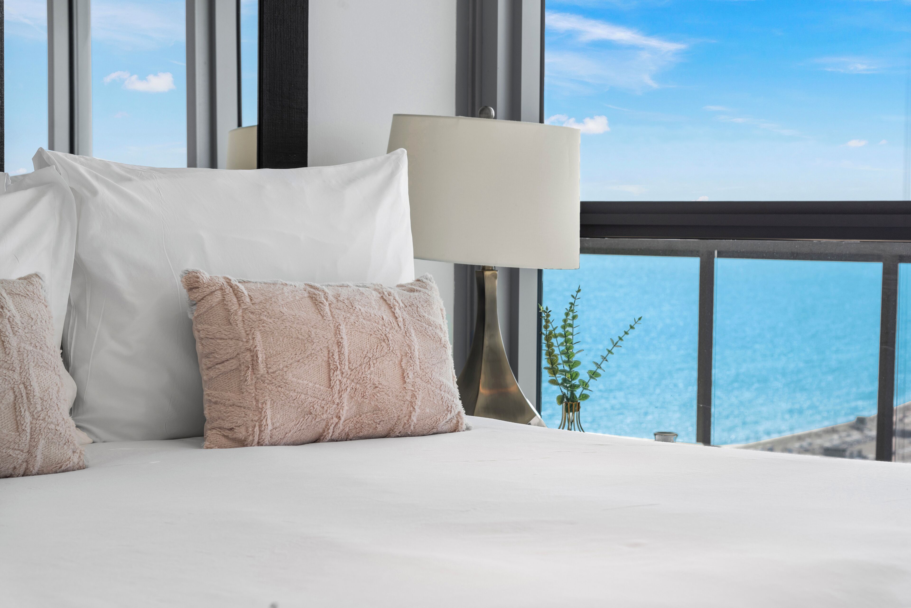 A bed with white and beige pillows, a bedside lamp, and a view of the ocean through large windows.