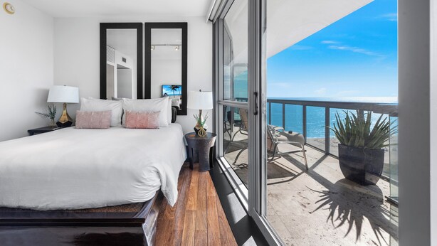 A bedroom with a large bed, bedside tables, a TV, and a balcony with a view of the sea.