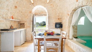 A rustic stone interior with a dining table, chairs, a small kitchen area, and a view of a swimming pool. - Trulli Salsapariglia by Perle di Puglia (Putignano)