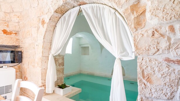 A stone-walled room with a clear blue pool, white curtains, and a white chair. - Trulli Salsapariglia by Perle di Puglia (Putignano)