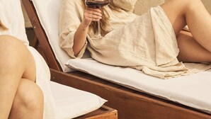 Eine Person lehnt auf einem hölzernen Liegestuhl mit einem weißen Kissen zurück und hält ein Glas Rotwein.