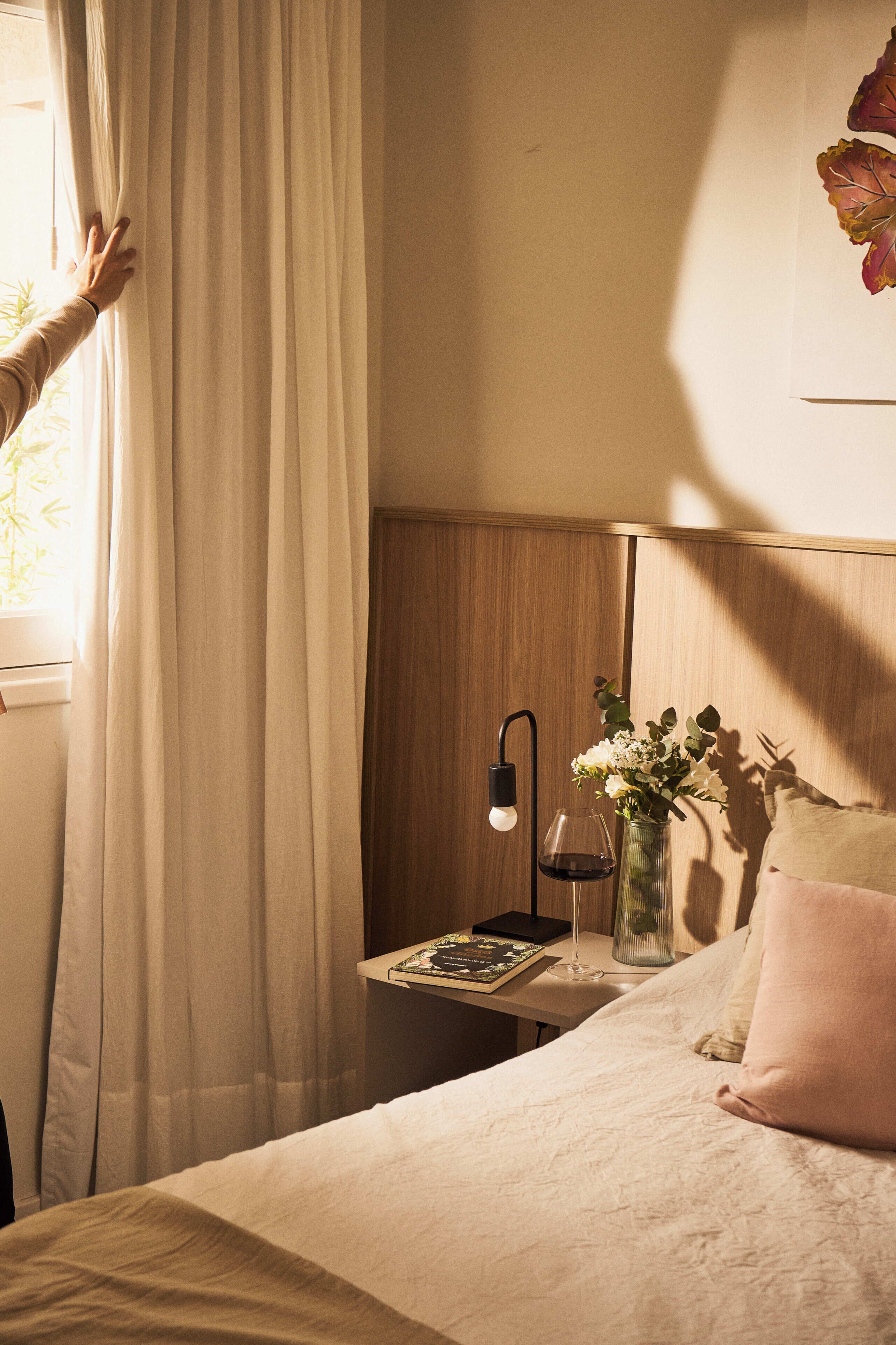 A bedroom with a bed, a bedside table with a lamp, a vase with flowers, and a window with curtains.