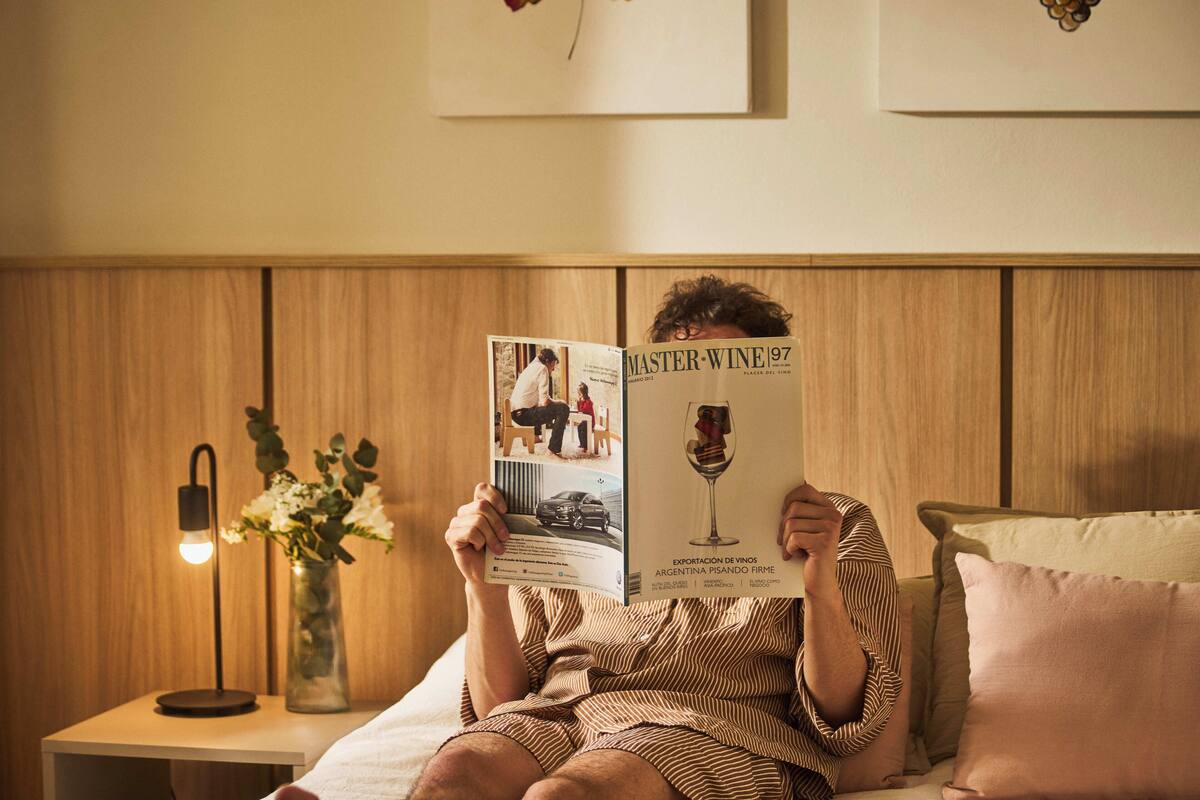 Iemand leest een tijdschrift met de titel 'Master Wine 97' in een kamer met een houten paneelwand, een nachtkastje met een lamp en een vaas met bloemen.