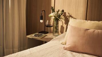 A neatly made bed with a white comforter and a single pink pillow. A bedside table holds a book, a glass of wine, and a vase with flowers.