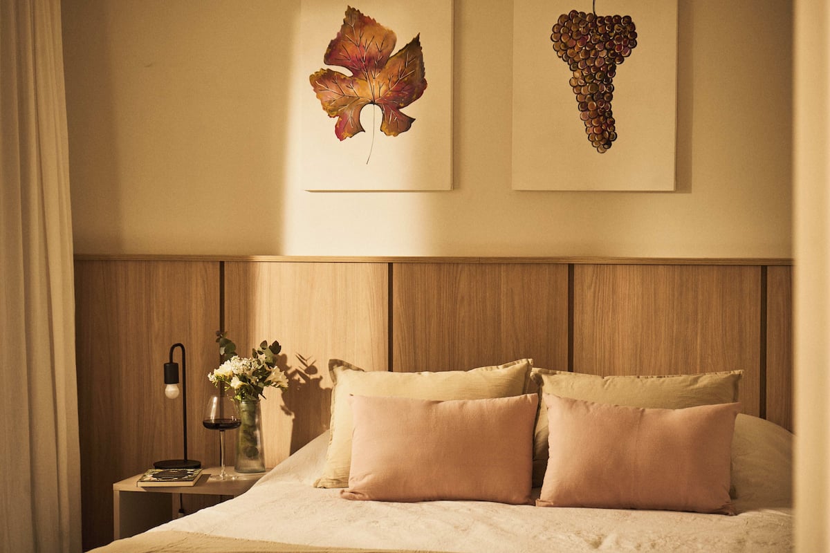 Een net opgemaakt bed met twee kussens, een nachtkastje met een bloemenvaas en een wijnglas, en twee kunstwerken boven het bed.