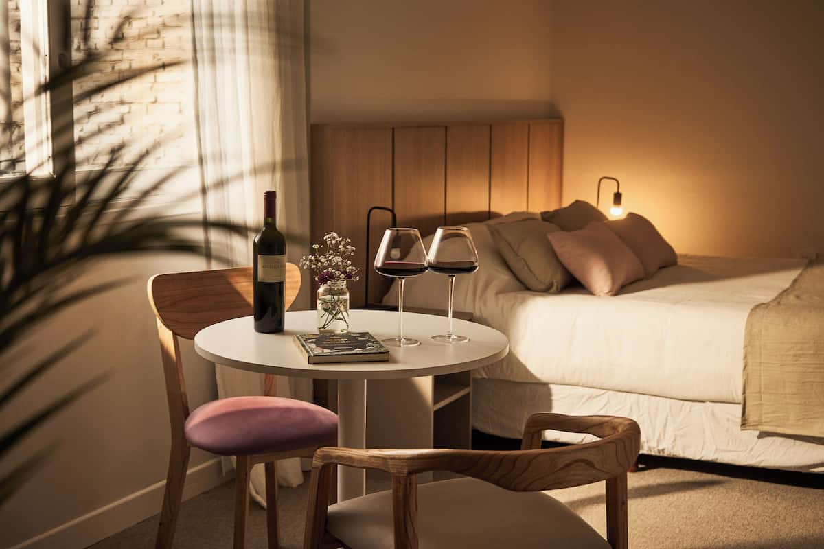 Een gezellige hotelkamer met een bed, een nachtkastje met twee wijnglazen, een fles wijn en een kleine vaas met bloemen.