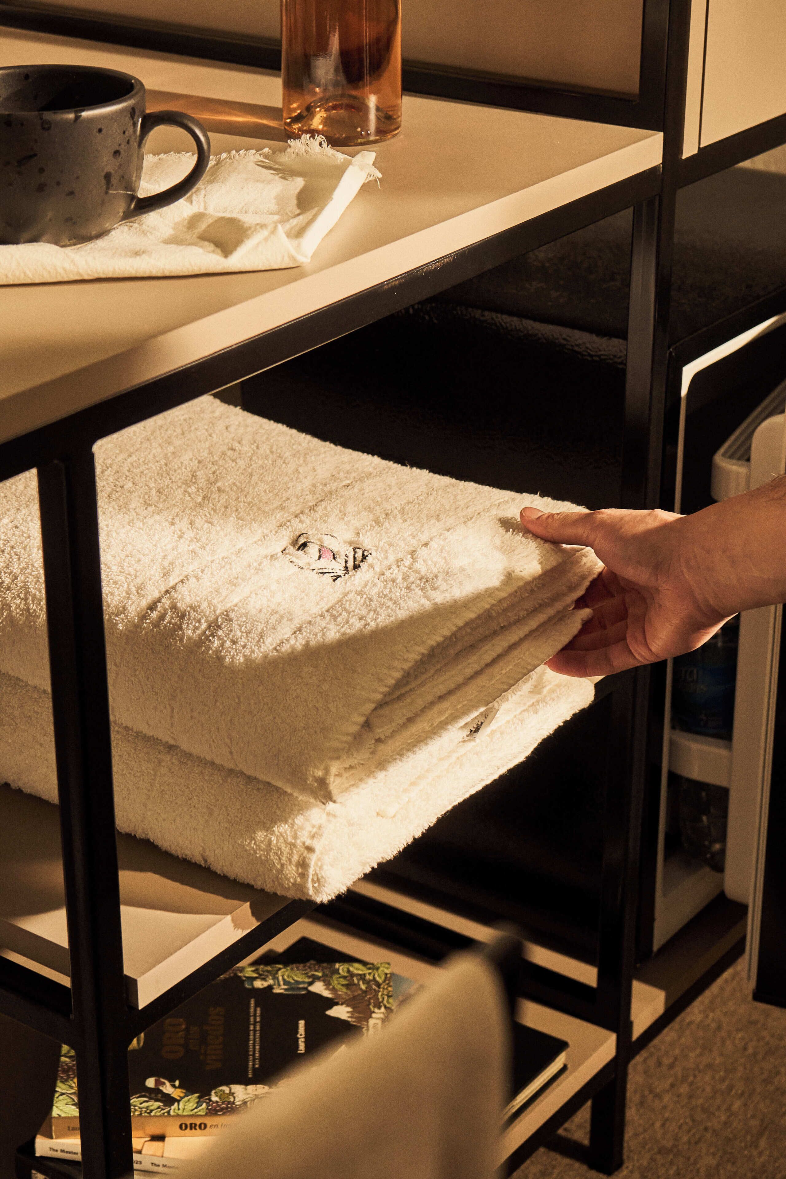 A hand reaching for a stack of white towels on a shelf in a room with a glass door and a framed picture on the wall.