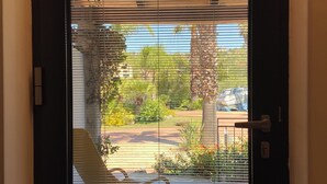 Blick durch ein Fenster mit horizontalen Rollläden, ein Stuhl und ein Garten mit Bäumen und Pflanzen.