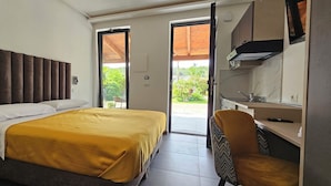 Ein Schlafzimmer mit einem Bett, einem Schreibtisch und einem Stuhl. Eine Tür führt zu einem Außenbereich mit Grünflächen.