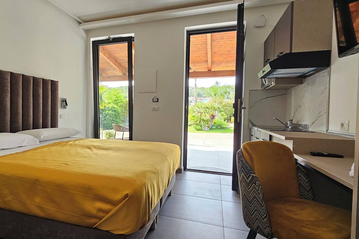 Ein Schlafzimmer mit einem Bett, einem Schreibtisch und einem Stuhl. Eine Tür führt zu einem Außenbereich mit Grünflächen.